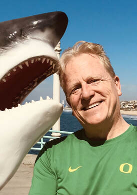 PHOTOGRAPH OF PORTLAND OREGON SCREENWRITER CHRIS MURRAY WITH BROAD SMILE PRETENDING THAT HE'S BEING ATTACKED BY FAKE SHARK IN OREGON DUCKS GREEN T-SHIRT