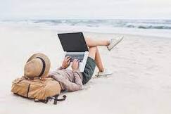 writer wearing hat, shirt, and shorts, reclining on a beach typing on laptop