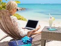 blonde young woman sitting in beach chair on white sand beach with laptop open on her lap and cocktail at her side