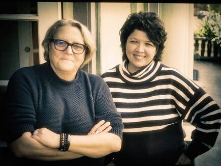 Photo of two women waist up, one blonde with short bobbed hair and wearing glasses with her arms folded across her chest, the other one on the right with short cropped hair and a nice smile wearing a black-and-white horizontally-striped sweater.